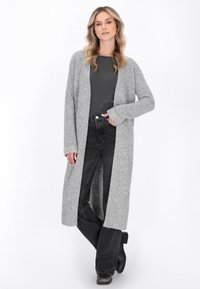 Donna che indossa un lungo cardigan grigio a maglia sopra una maglietta grigio scuro, jeans neri e stivali neri, in piedi con una mano in una tasca.
