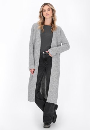 Femeie îmbrăcată cu o cardigan lung, gri, din tricot, peste un top gri închis, blugi negri și cizme negre, stând cu o mână în buzunar.