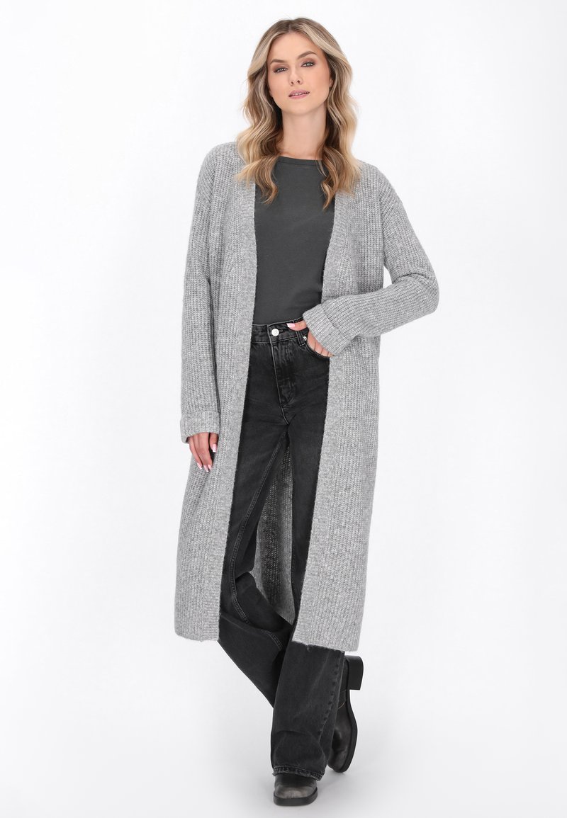Donna che indossa un lungo cardigan grigio a maglia sopra una maglietta grigio scuro, jeans neri e stivali neri, in piedi con una mano in una tasca.