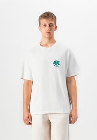 Ung mand iført hvid t-shirt med grønt abstrakt logo på brystet og lyse bukser, stående foran en ensfarvet hvid baggrund.