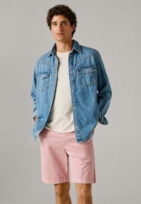 Nuori mies seisoo päällään vaaleansininen farkkupaita valkoisen T-paidan päällä ja vaaleanpunaiset polvenmittaiset shortsit harmaan yksivärisen taustan edessä.