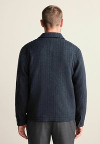 Donkerblauwe wolblendjas met een herringbone textuur, gestructureerde kraag en knoopmanchetten. Rechte snit met een schone achterkant.