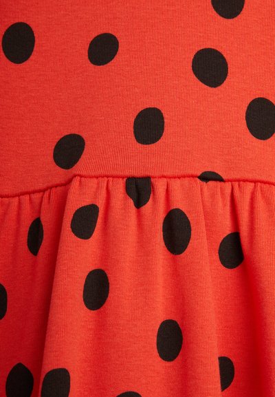 Tissu rouge à pois noirs. Le design présente une couture froncée à la taille, créant un effet volanté près du bas.