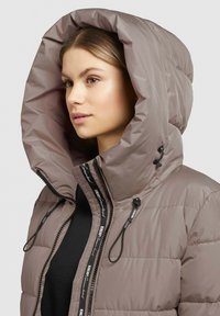 Giacca a vento imbottita in marrone chiaro con un ampio cappuccio. Le caratteristiche includono una chiusura a zip sul davanti, cordini e una texture trapuntata. Accenti vicino alla zip.