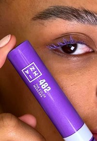 Purple mascara tube labeled "ZW 482 THE COLOR MASCARA" held near an eye with matching purple mascara applied to lashes. Smooth, glossy finish.