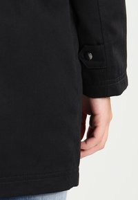 Svart bomullsjacka med en knappknäppning på ärmen, som visar tygtextur och sömnadsdetaljer. Hälften av jackan hålls i handen.