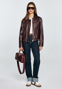 Bordeauxfarbene Lederjacke mit zwei großen Taschen, kombiniert mit dunklen Jeans, einer strukturierten weinroten Tasche und weiß-bordeauxfarbenen Sneakers.