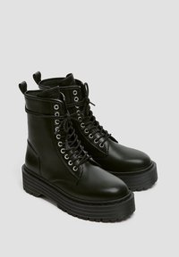 Bottes de combat en cuir noir avec bout rond, semelle plateforme épaisse et œillets en métal. Caractérisées par un design à lacets et des languettes de tirage à la cheville.