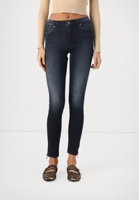 Frau trägt dunkelblaue Skinny-Jeans mit hoher Taille und Reißverschlussdetails an den Knöcheln sowie Leopardenmuster-Ballerinas mit schwarzen Riemen.
