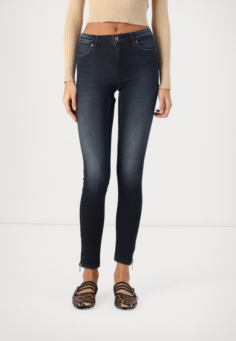 Frau trägt dunkelblaue Skinny-Jeans mit hoher Taille und Reißverschlussdetails an den Knöcheln sowie Leopardenmuster-Ballerinas mit schwarzen Riemen.