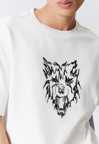 Bílý krátký rukáv pánský tričko z bavlny s černým grafickým potiskem stylizované hlavy vlka na hrudi. Hladká textura, kulatý výstřih.