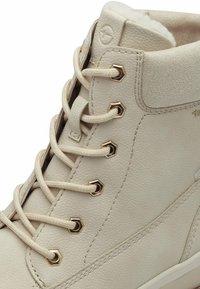Bottines en cuir beige avec des œillets en or hexagonaux, des lacets plats et une doublure douce. Présente une texture subtile et des accents cousus.