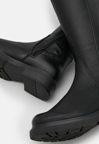 Las botas de cuero negro hasta la rodilla cuentan con un cierre lateral, suela de goma plana y parte superior texturizada, mostrando un diseño elegante con acentos mínimos.