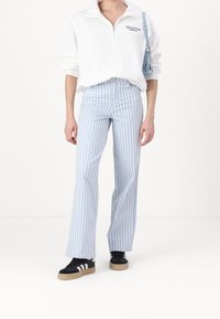 Vit fleecejacka med dragkedja och logotyp, kombinerad med ljusblå vertikalt randiga utsvängda byxor och svarta sneakers med vita accenter.