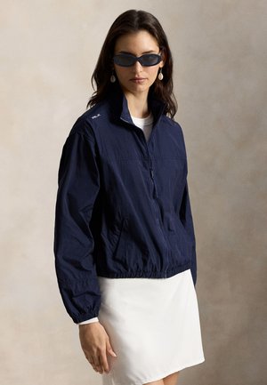 Μπλε ναυτικό ζακέτα με φερμουάρ από ελαφρύ ύφασμα, με ψηλό γιακά, ελαστική μέση και δύο πλαϊνές τσέπες· κεντημένη λεπτομέρεια λογότυπου.