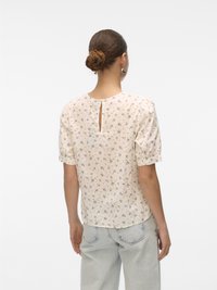 Floral, short-sleeve top in cream, featuring pastel flower patterns, a back keyhole detail, and relaxed fit. Paired with light wash jeans.