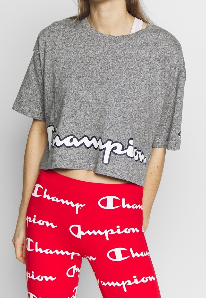 Grå kortärmad t-shirt med en framträdande vit och marinblå "Champion"-logotyp längs fållen, bärs med röda leggings märkta med samma logotyp.
