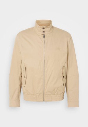Beige lättviktsjacka med hög krage, frontdragkedja, sidofickor och elastiska ärmslut. Texturerad tyg med en subtil lyster.