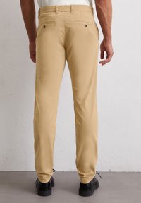 Beżowe chinosy o slim fit, z dwiema tylne kieszeniami z guzikami, szlufkami na pasek i gładką teksturą bawełnianą. Czarne buty dopełniają całość.