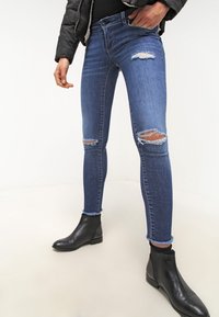 Denim jeans in dark blue with a skinny fit, featuring distressed details and frayed cuffs, paired with black ankle boots.