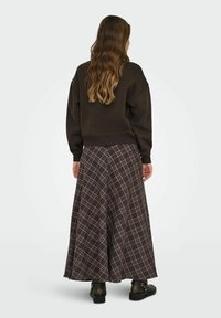 Sudadera marrón con hombros caídos combinada con una falda larga de cuadros que presenta líneas marrones, blancas y lilas claras, usada con botas negras.