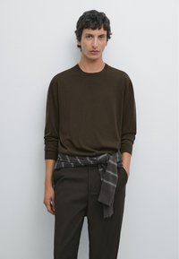 Brauner Pullover mit langen Ärmeln und rundem Ausschnitt, kombiniert mit dunkler Hose und einem grauen, karierten Schal, der in der Taille gebunden ist. Schlichter Hintergrund.