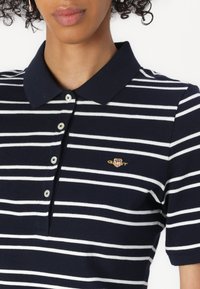 Námořnická modrá polo tričko s bílými pruhy, s klasickým límcem a zapínáním na knoflíky. Na hrudi je umístěno malé zlaté logo.