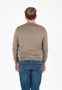 Uomo con capelli corti castano chiaro, vestito con un maglione beige e jeans blu, di spalle su uno sfondo bianco.