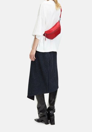 Rode leren schoudertas met een gebogen vorm en ritssluiting, gedragen over een witte oversized T-shirt en een marineblauwe midi-rok met strepen. Zwarte kniehoge laarzen.