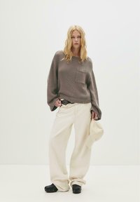 Frau mit lockigem blonden Haar, die einen braunen oversized Pullover und weite cremefarbene Hosen sowie schwarze Clogs trägt. Sie hält eine beige Mütze vor einem weißen Hintergrund.