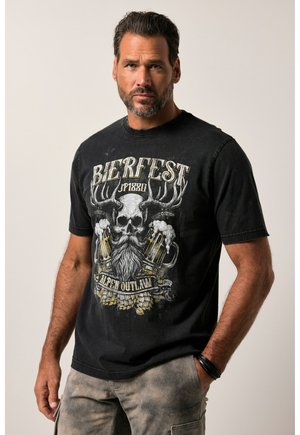 Mand med skæg iført sort T-shirt med kranium, gevirer, ølkrus og teksten "Bierfest Alpen Outlaw", stående med den ene hånd i lommen.