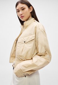 Jauna sieviete, ģērbta bēšā īsā žaketē ar priekšējām plakanajām kabatām un baltās biksēs, pozē pret vienkāršu gaišu fonu.