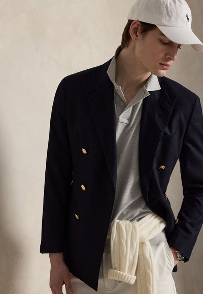 Νέος άνδρας που φοράει λευκό καπέλο, μπλε σακάκι με χρυσά κουμπιά, γκρι πόλο πουκάμισο, εκρού πουλόβερ δεμένο στη μέση και λευκό παντελόνι, ποζάρει μπροστά σε μπεζ φόντο.