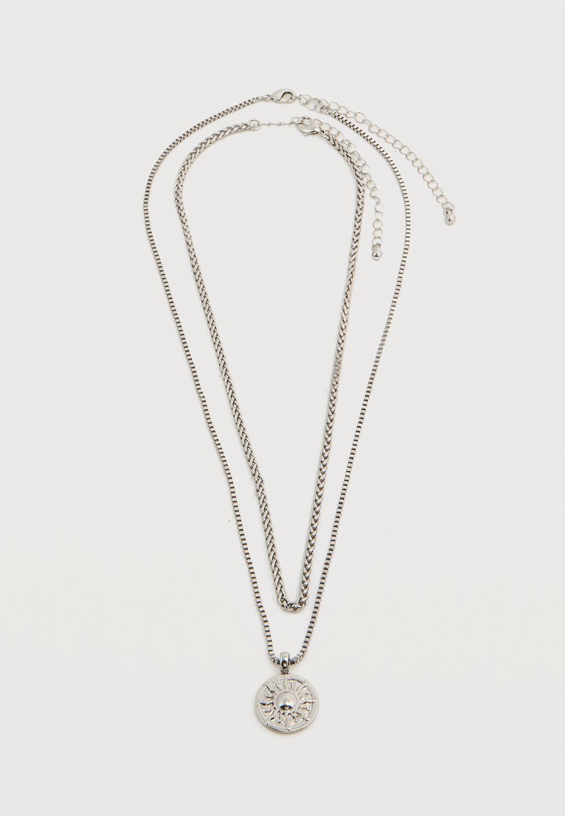 Δύο ασημί κολιέ σε στρώσεις, το ένα με στρογγυλό μενταγιόν με μοτίβο ήλιου, και τα δύο με ρυθμιζόμενα κούμπωμα αλυσίδας σε λευκό φόντο.