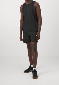 Hombre con camiseta atlética sin mangas negra, pantalones cortos deportivos negros con logotipo amarillo, calcetines blancos y zapatillas deportivas negras de pie sobre un suelo gris.