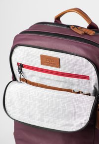 Zaino marrone con una texture liscia, dotato di un'etichetta in pelle marrone e di una tasca con zip rossa. L'interno presenta un motivo a griglia e tasche aggiuntive.