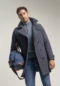 Abrigo gris estilo trench con grandes botones, sobre un suéter tejido en azul claro con relieve de cables. Vaqueros y un bolso tipo duffle azul con detalles en marrón.