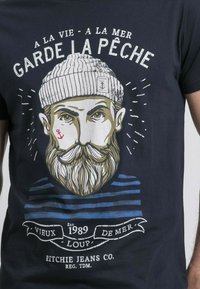 T-shirt bleu marine avec un motif d'homme barbu portant un bonnet en tricot. Texte en gras au-dessus et rayures en dessous de l'illustration. Matière en coton.