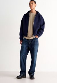 Veste zippée à capuche bleue marine, t-shirt beige et jeans bleu foncé. L'ensemble présente des couleurs unies et une coupe décontractée, avec des bottes noires.