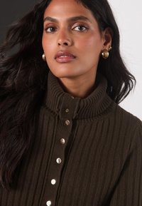 Cardigan marrone a coste con collo alto, caratterizzato da bottoni tono oro sul davanti. Tessuto morbido con design casual e senza motivi visibili.