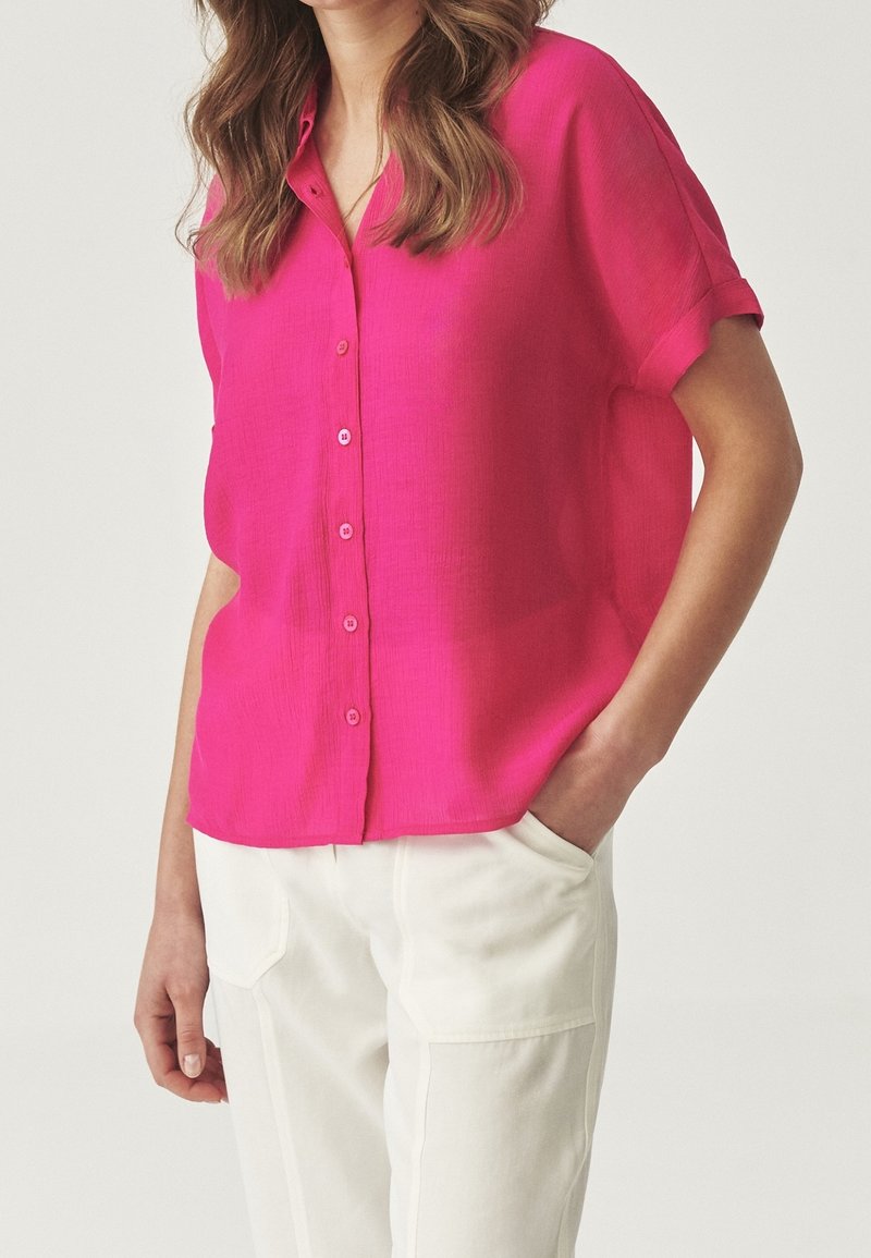 Blusa rosa a maniche corte con design a bottoni sul davanti e vestibilità rilassata. Realizzata in tessuto leggero, presenta una texture sottile e polsini arrotolati.