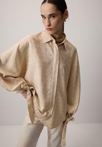 Bluză bej cu mânecă lungă, cu un finisaj lucios, având modele circulare embosate, cu închidere cu nasturi și manșete legate. Asortată cu pantaloni crem.