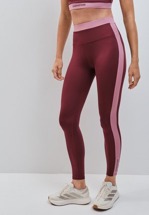 Frau trägt bordeauxrote Leggings mit pinken Seitenstreifen und weißen Sneakers, steht vor einem einfarbigen hellgrauen Hintergrund.