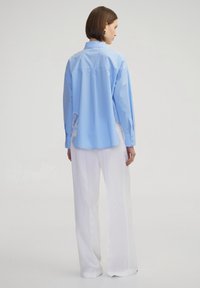 Lichtblauw overhemd met lange mouwen, voorzien van een gehaakt kantaccent aan de zoom, gecombineerd met witte wijde broek. Glad stofafwerking.