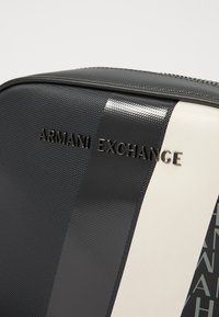 Svart og hvit reisekoffert med teksturert overflate, med en fremtredende "Armani Exchange"-logo og strømlinjeformede designdetaljer.