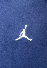 Tecido azul com um logótipo Jumpman bordado em branco, que representa uma figura na posição de salto. A textura parece suave e semelhante a fleece.