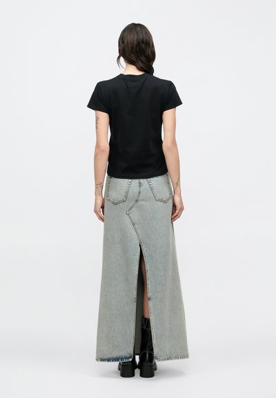 Žena s dlhými tmavými vlasmi nosí čierne tričko s krátkym rukávom, dlhú svetlú rifľovú sukňu s rozparkom vzadu a čierne členkové čižmy, otočená chrbtom.