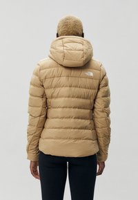 Chaqueta acolchada beige con un diseño ajustado, capucha y textura acanalada. Cuenta con un logotipo en la parte superior de la espalda y un patrón de acolchado horizontal.