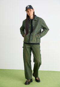 Jachetă din denim verde și pantaloni cargo, cămașă neagră dedesubt, șapcă neagră și pantofi cu model. Design simplu, croială lejeră, detalii de cusătură vizibile.