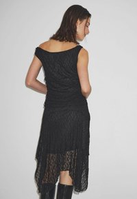 Fekete csipkeből készült ruha aszimmetrikus szegéllyel, testhez simuló felsőrésszel és váll nélküli kivágással. A textúrált anyag és a réteges szoknya részlet fokozza az eleganciát.